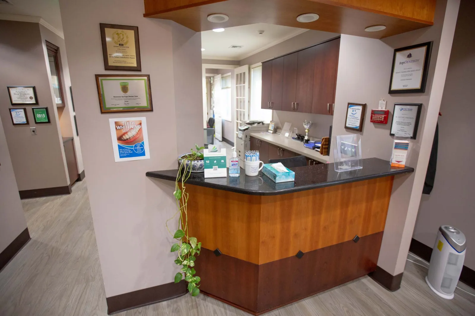 Reception desk area with wood paneling, granite countertop, certificates on beige walls, and office supplies in dental clinic.