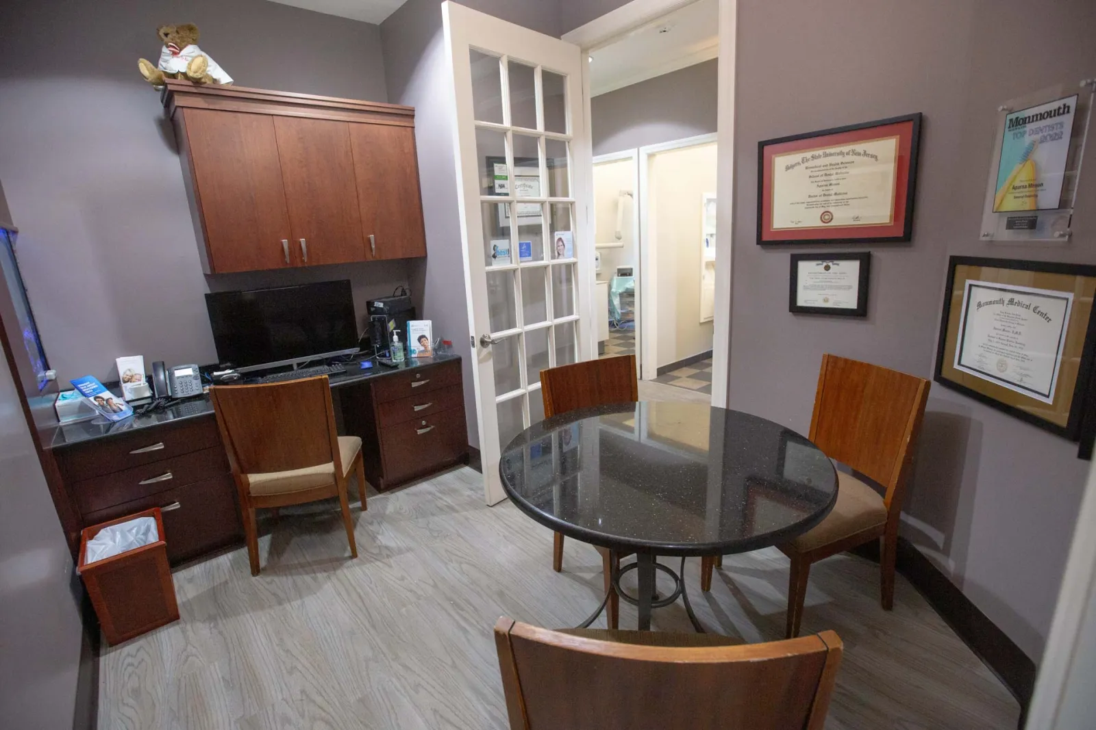 Small professional office with round table, three chairs, computer desk, certificates on gray walls, and wood cabinetry.