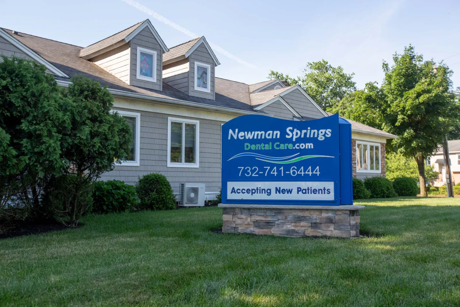 Newman Springs Dental Care office building with a blue sign accepting new patients outside on a grassy lawn.