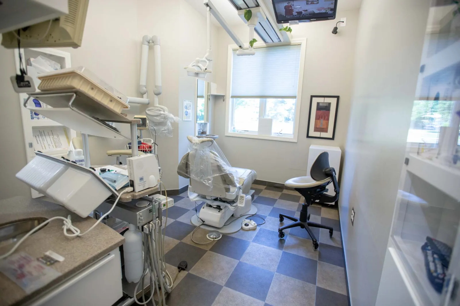 Bright dental office with patient chair, dental equipment, and a TV mounted near the window on checkered floor.