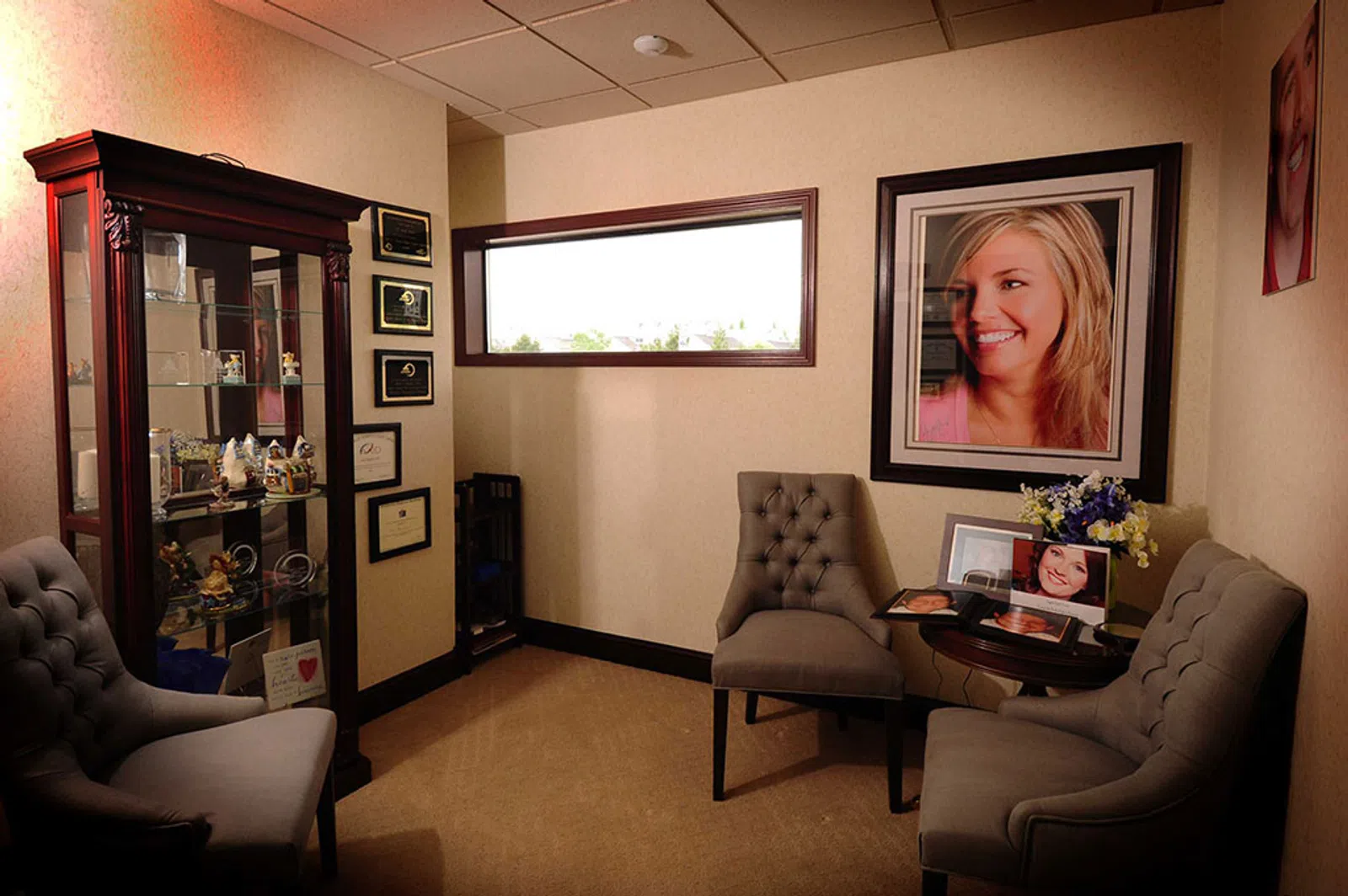 Cozy waiting room with tufted chairs, framed portraits, a glass display cabinet, and wall certificates.