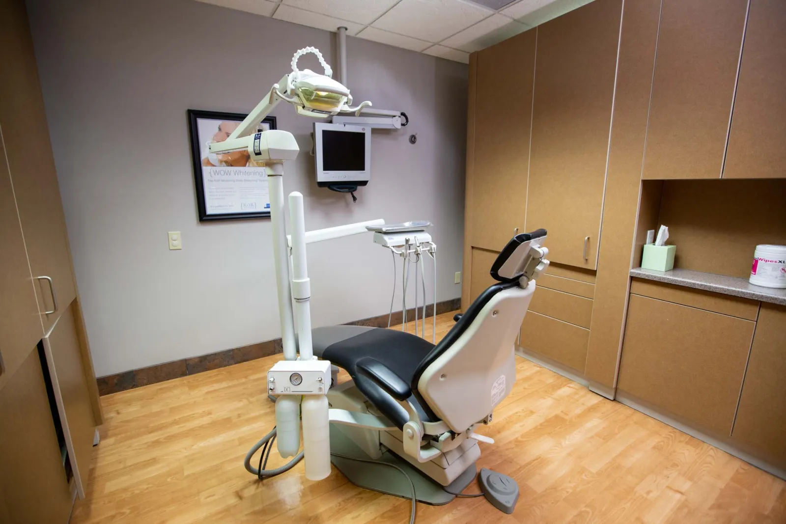 Modern dental clinic room with dental chair, overhead light, monitor, and cabinetry on wooden floor.