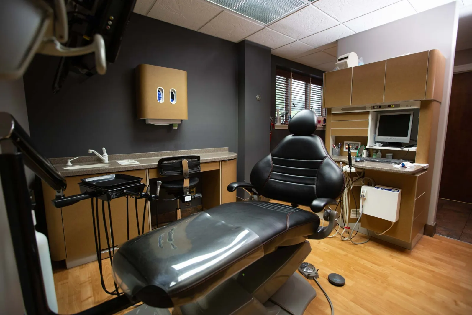 Modern dental office with black dental chair, cabinetry, computer monitor, and medical equipment on wood floor.