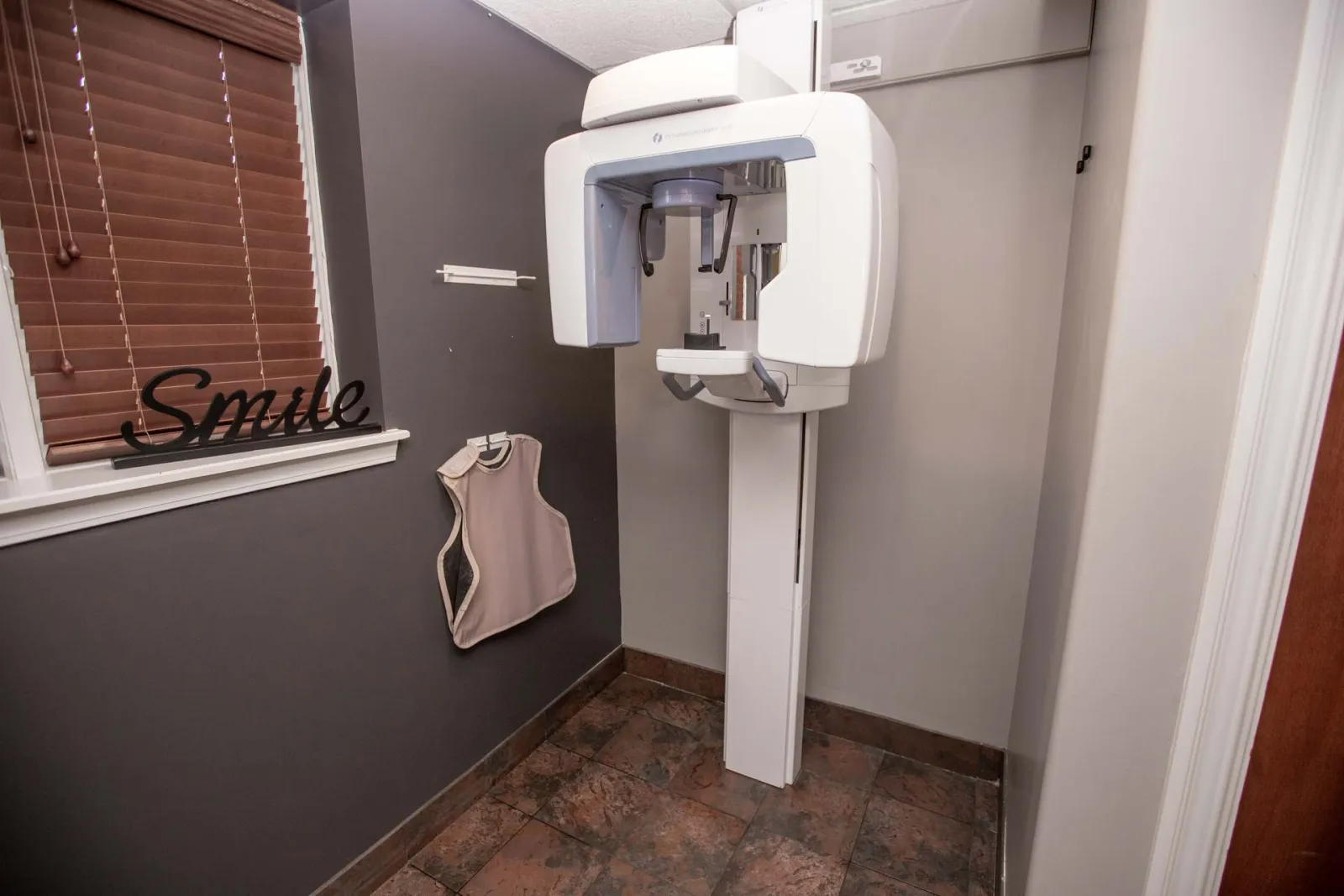 Dental X-ray machine in a small exam room with a protective apron hanging and a smile sign on the window.
