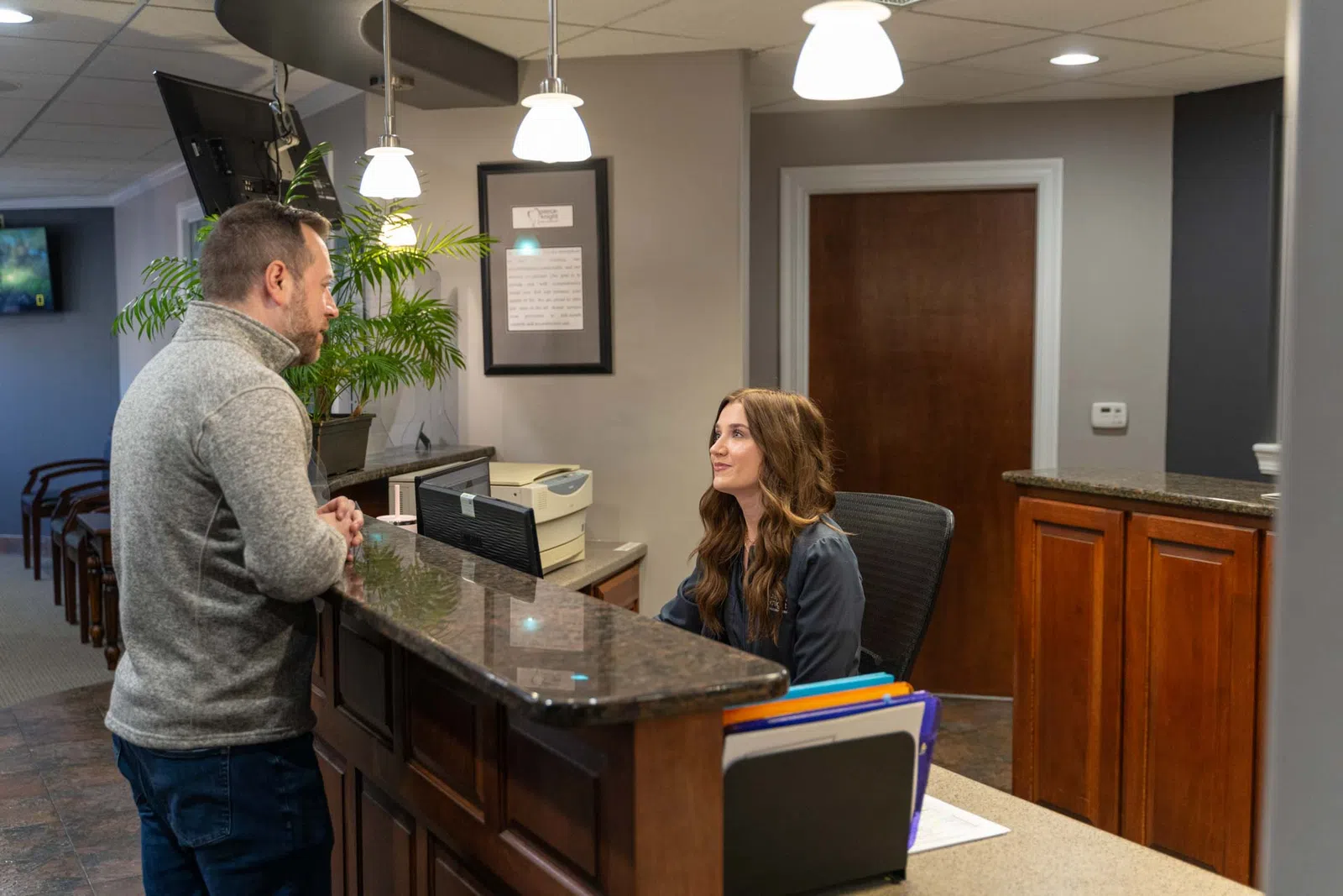 Man in gray sweater talking to receptionist at office front desk with computer and files