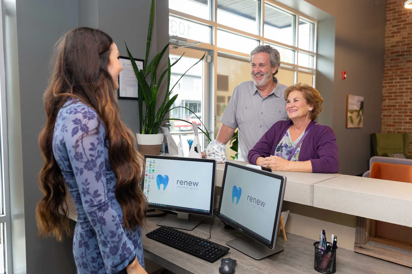 Dental clinic receptionist assisting elderly couple at front desk with computer screens displaying Renew Dentistry logo.