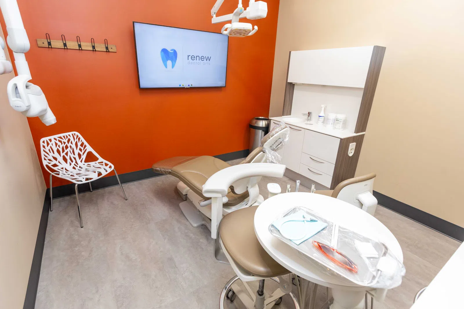 Modern dental office with beige chair, orange accent wall, white shelving, and screen displaying Renew Dental Arts logo.