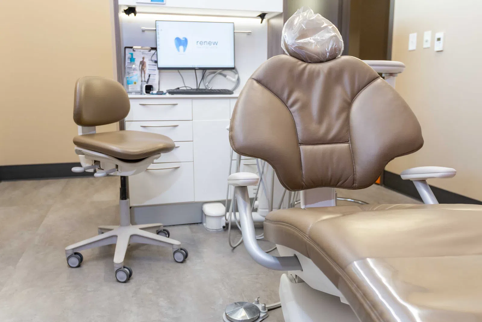 Modern dental clinic room with brown dental chair, dentist stool, and monitor displaying a tooth logo.