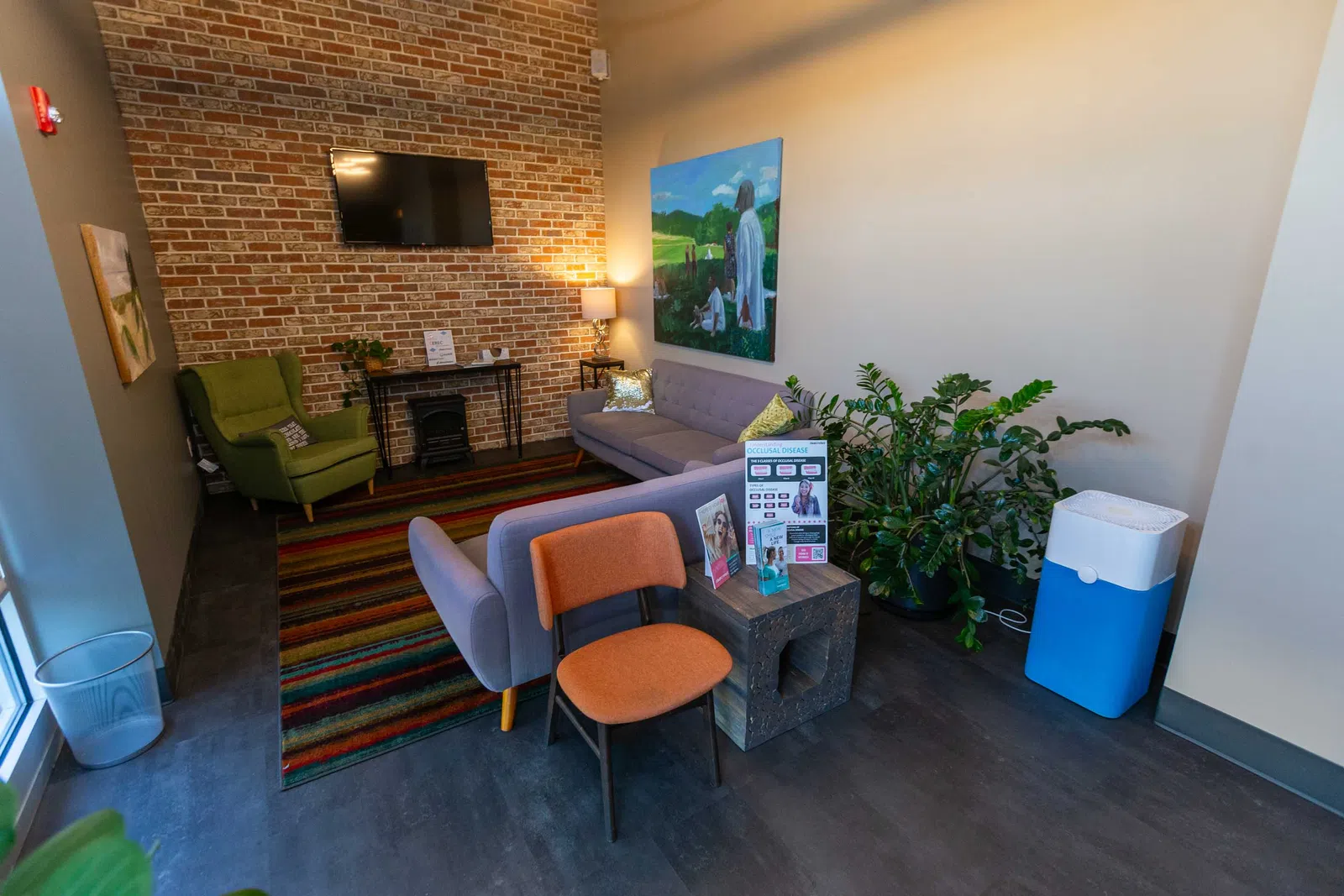 Cozy waiting room with brick wall, TV, green armchair, gray sofa, colorful rug, and indoor plants