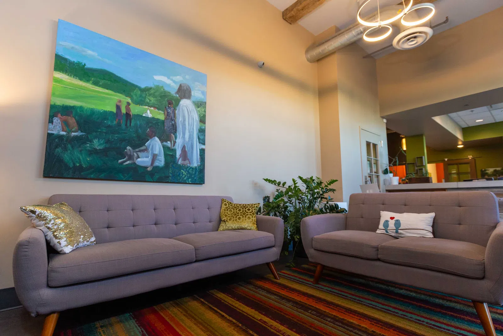 Modern living room with gray sofas, colorful rug, green plants, and a large landscape painting on the wall.