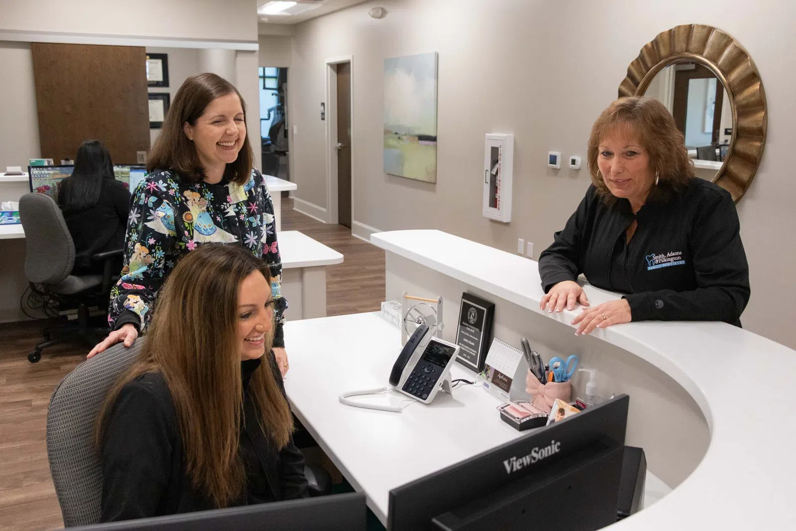 Three women smiling and working together at a reception desk inside a modern office space.