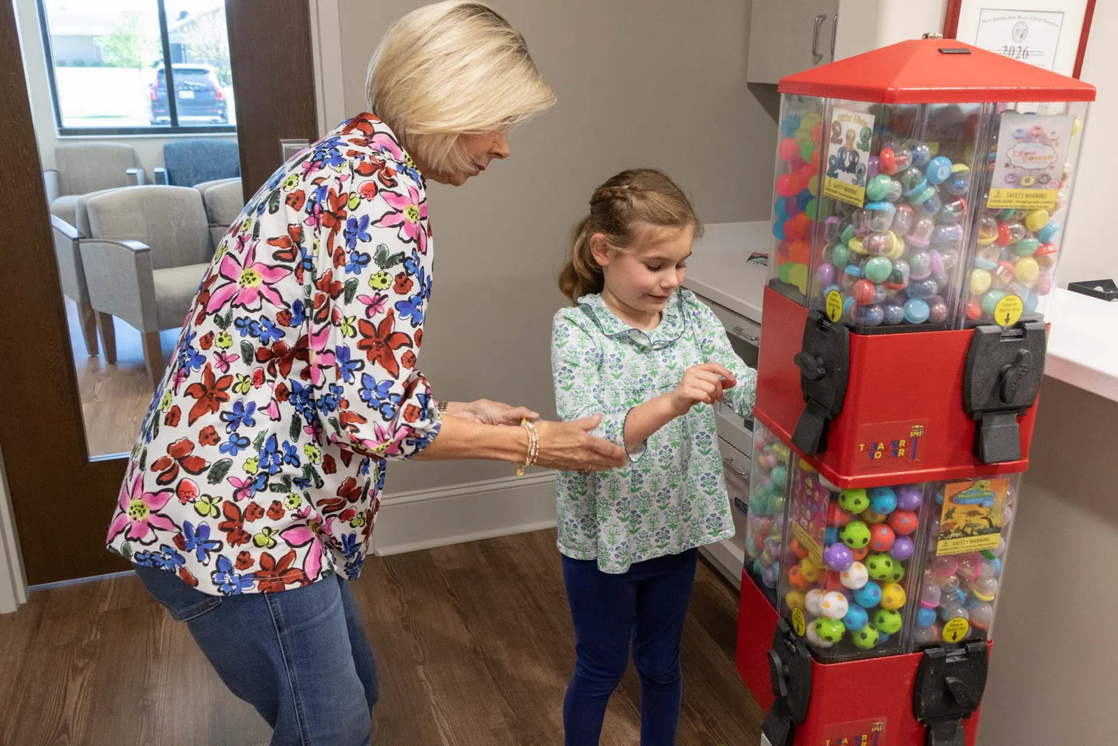 Child and woman using a red gumball machine filled with colorful balls in a bright room.
