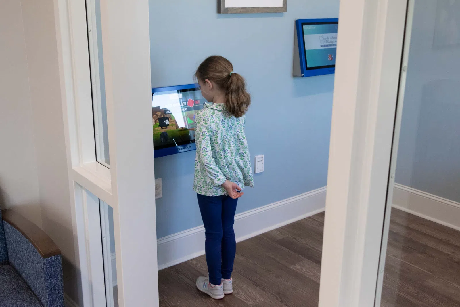 Young girl interacting with a touch screen display in a modern waiting room with blue walls and wooden floor.
