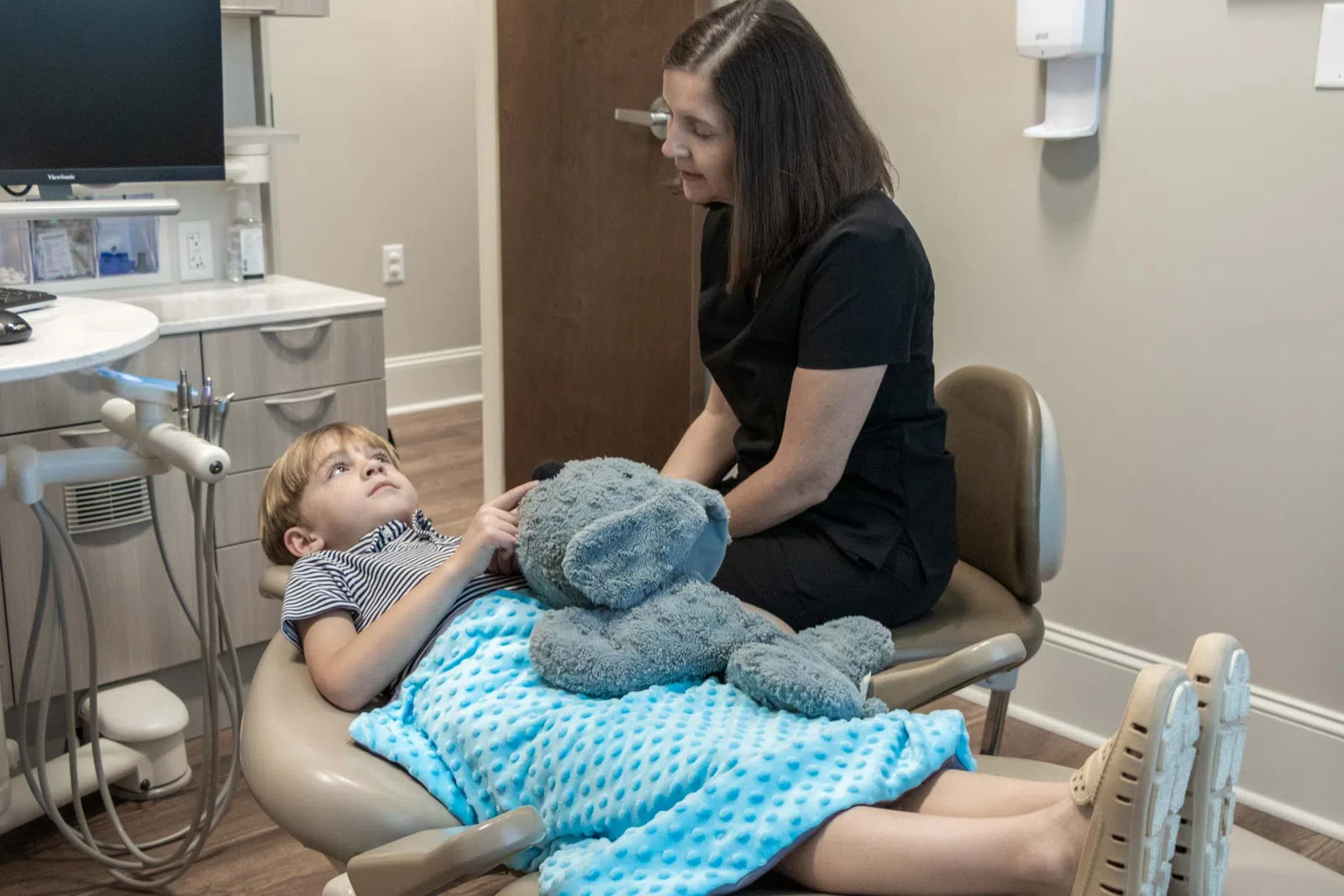 Child lying on dental chair covered with blue blanket holding teddy bear while dentist talks to him