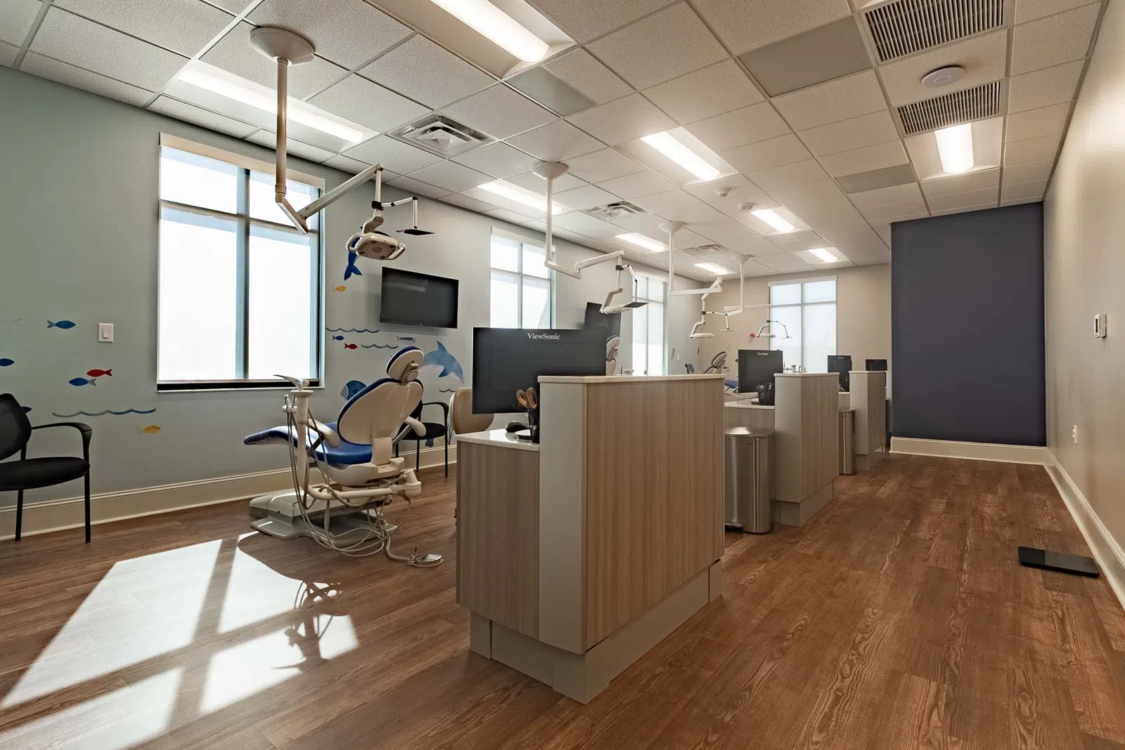 Modern dental clinic interior with treatment chairs, monitors, wood flooring, and ocean-themed wall decals.