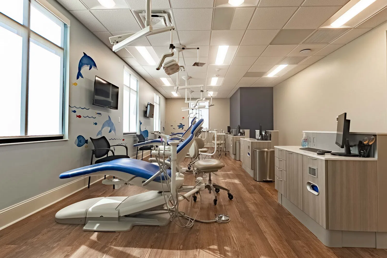 Bright dental office with blue chairs, marine-themed wall art, wood flooring, and modern dental equipment.