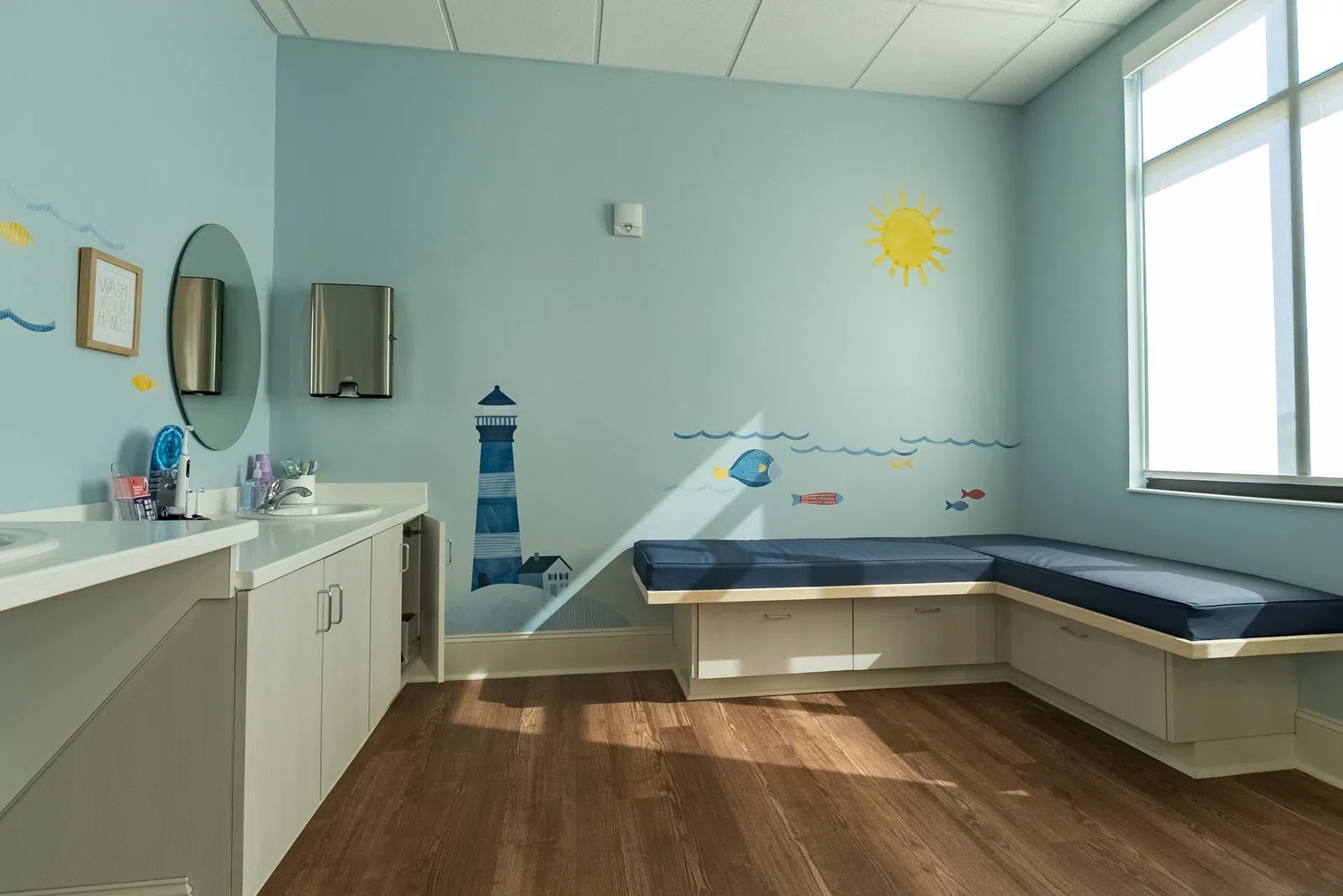 Bright children's clinic room with blue nautical wall art, bench seating, white cabinets, and large window letting in sunlight