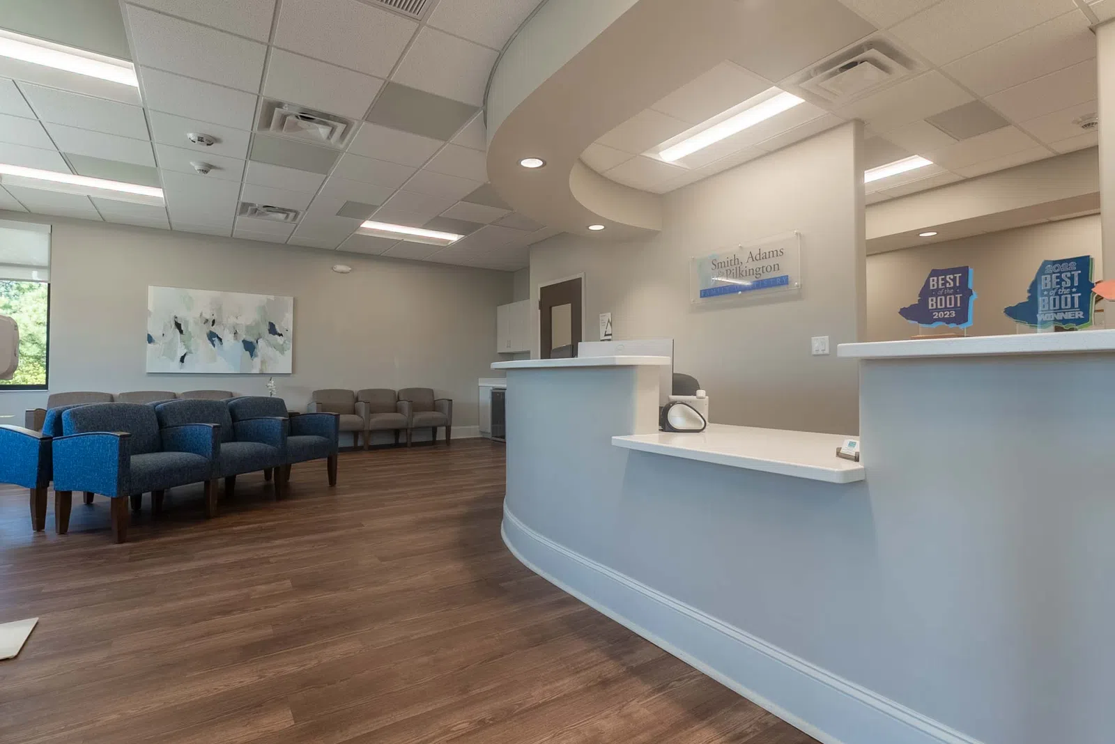 Modern medical office reception area with curved white desk, blue and gray chairs, and wood flooring.