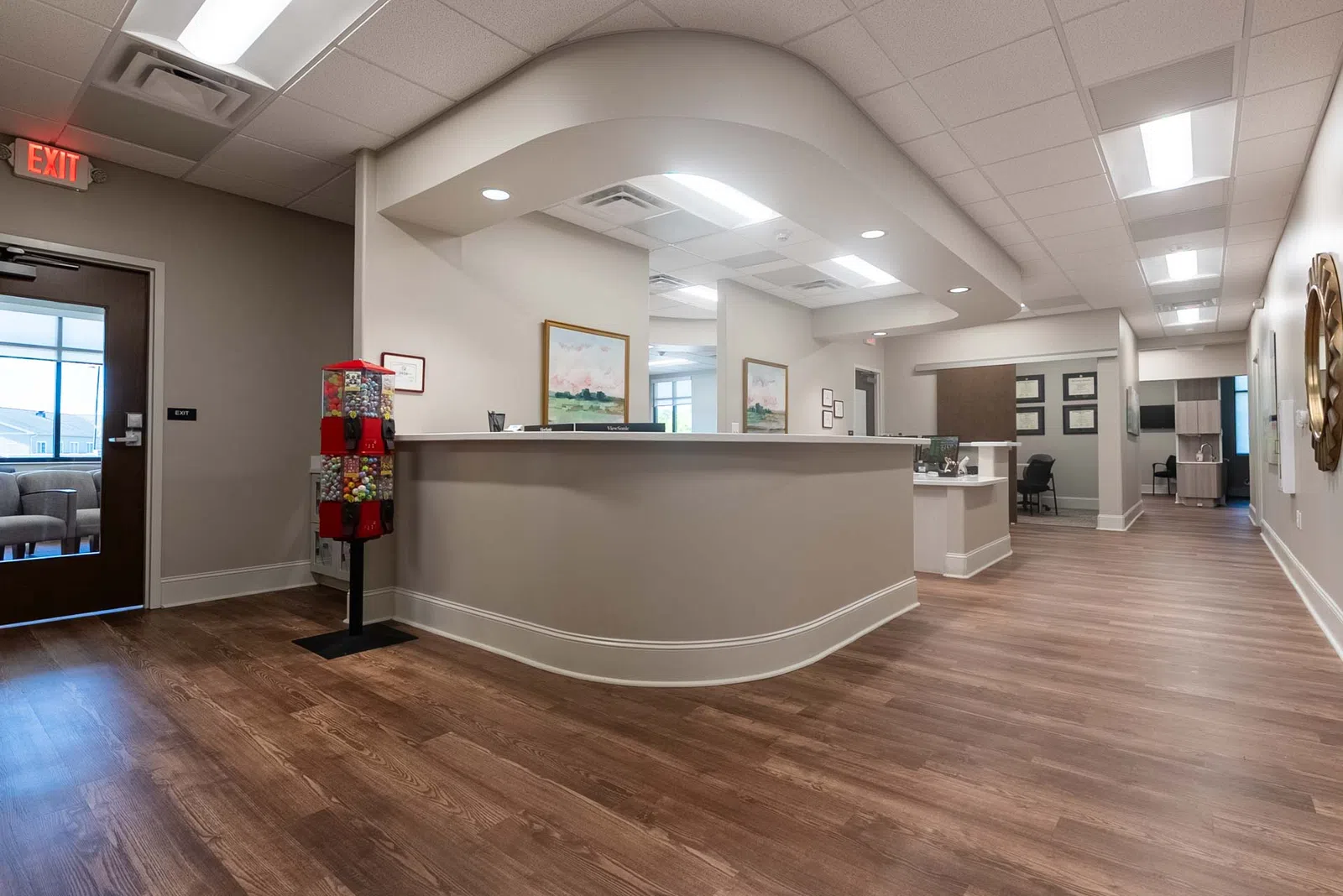 Modern office reception area with curved desk, wood flooring, gumball machine, and contemporary artwork on walls.