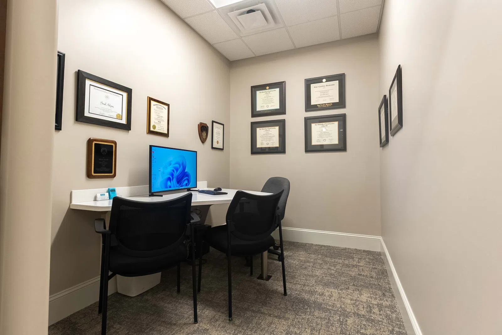 Small professional office with computer, two chairs, beige walls, and framed certificates on the walls.