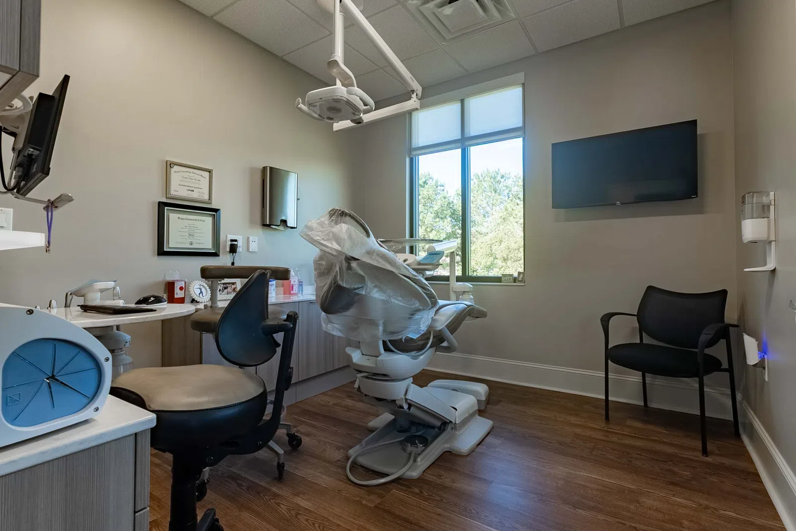 Modern dental office with patient chair, medical equipment, certificates, and large window with natural light.