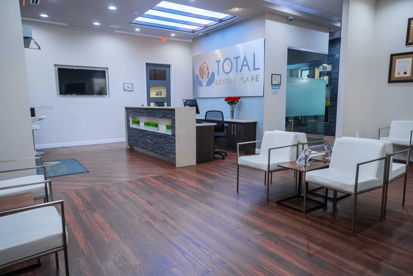Modern dental office waiting area with white chairs, wood flooring, reception desk, and Total Dental Care sign.
