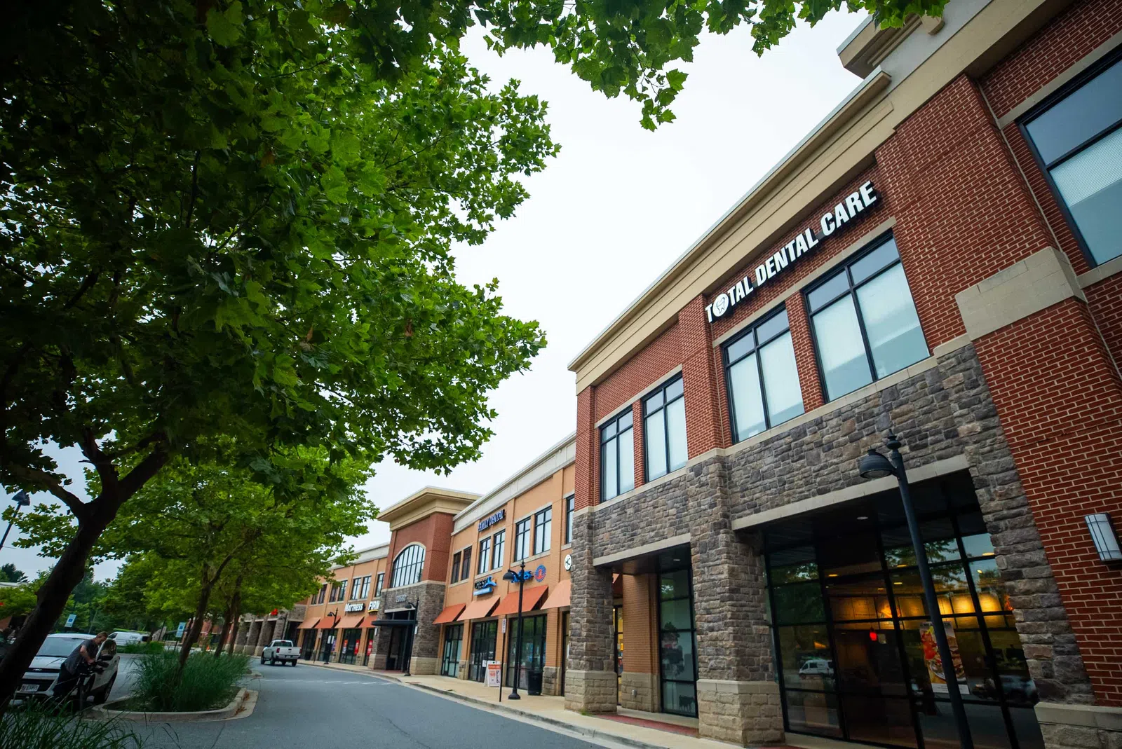 Modern commercial building with Total Dental Care and other storefronts under green trees on a cloudy day.