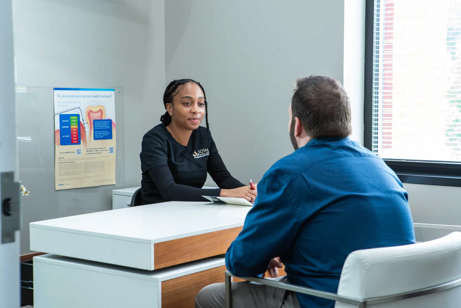 Female healthcare professional consulting a male patient in a modern office with dental health poster visible.