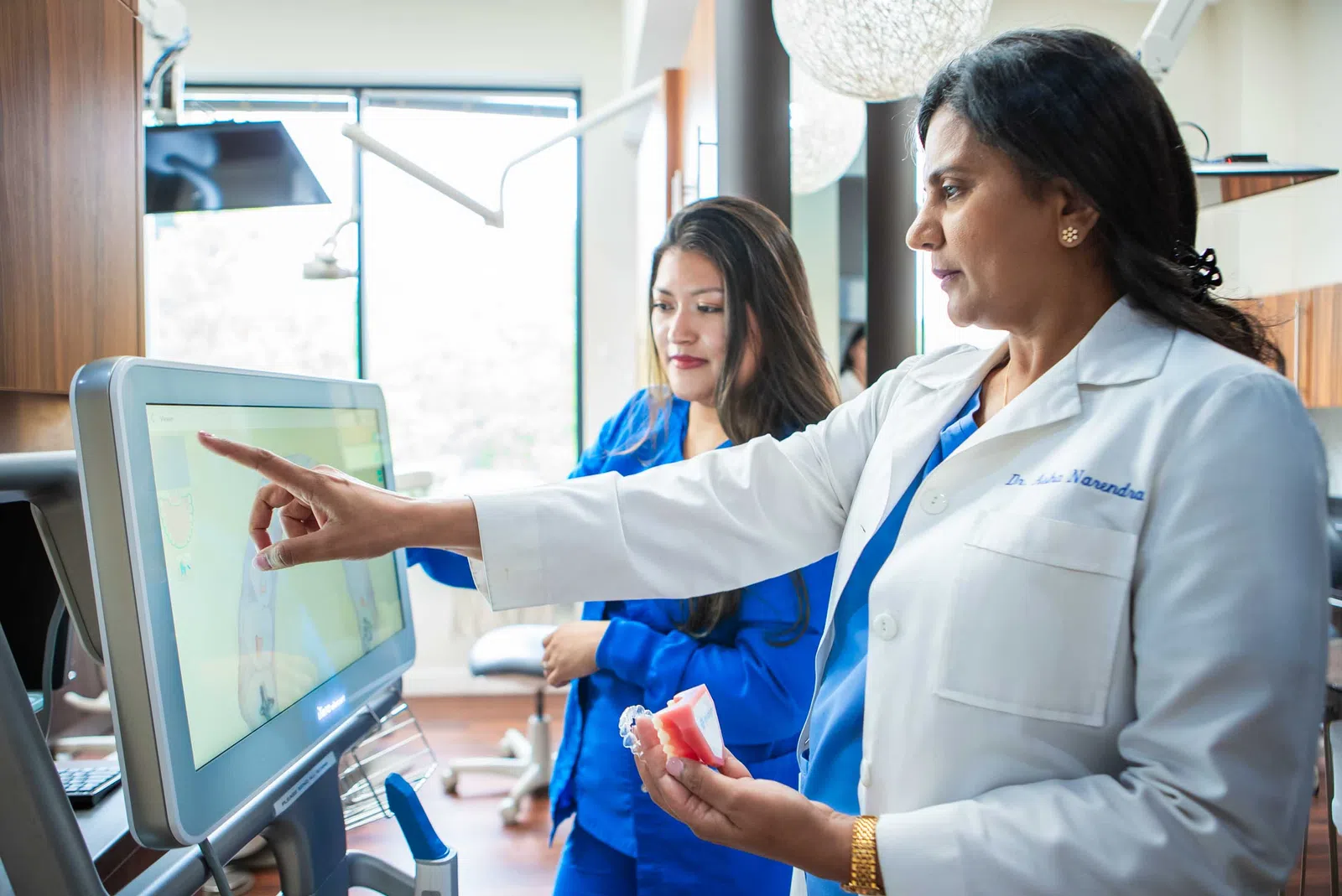 Female dentist in white coat showing dental model and pointing at screen with assistant in clinic.
