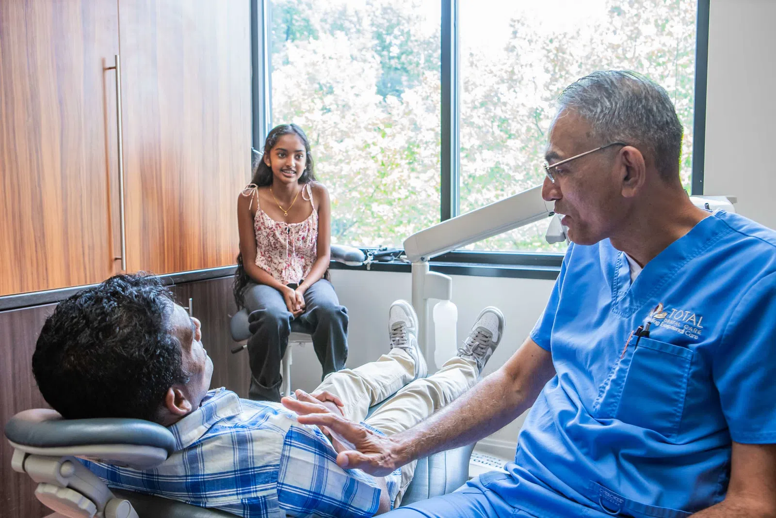 Dentist in blue scrubs talking to male patient in dental chair while young girl sits nearby by window.