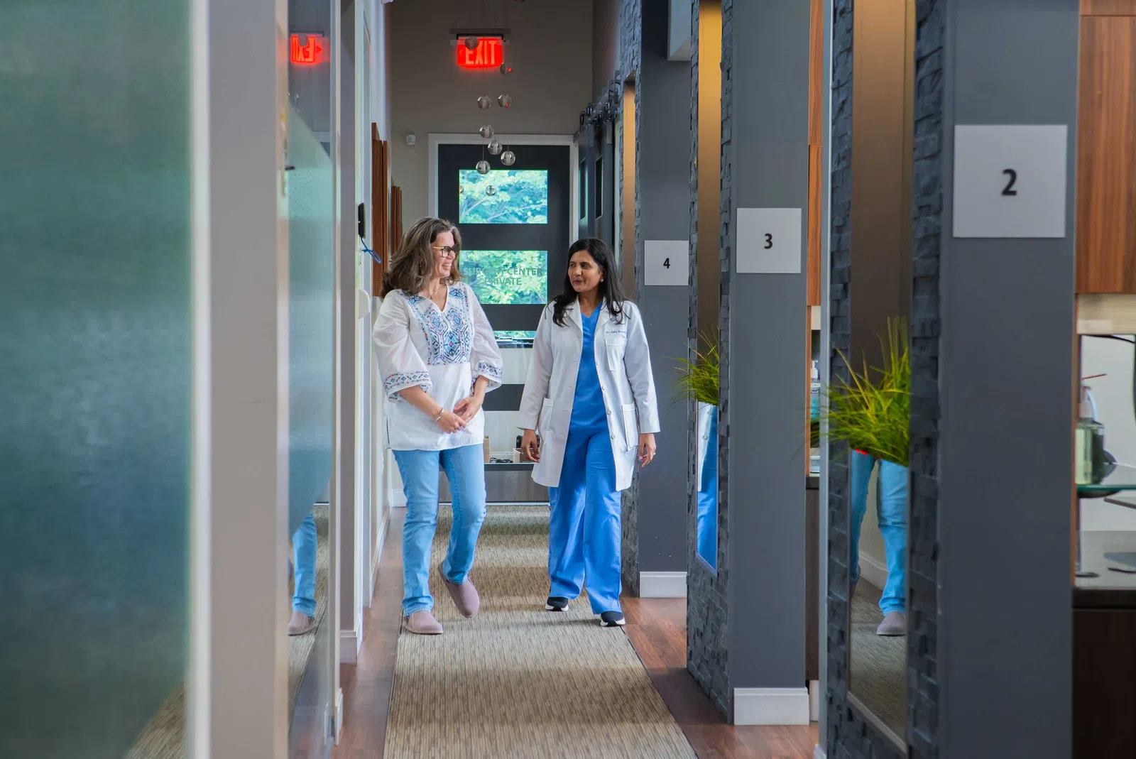Doctor and patient walking and talking together in a modern medical clinic hallway with numbered rooms.