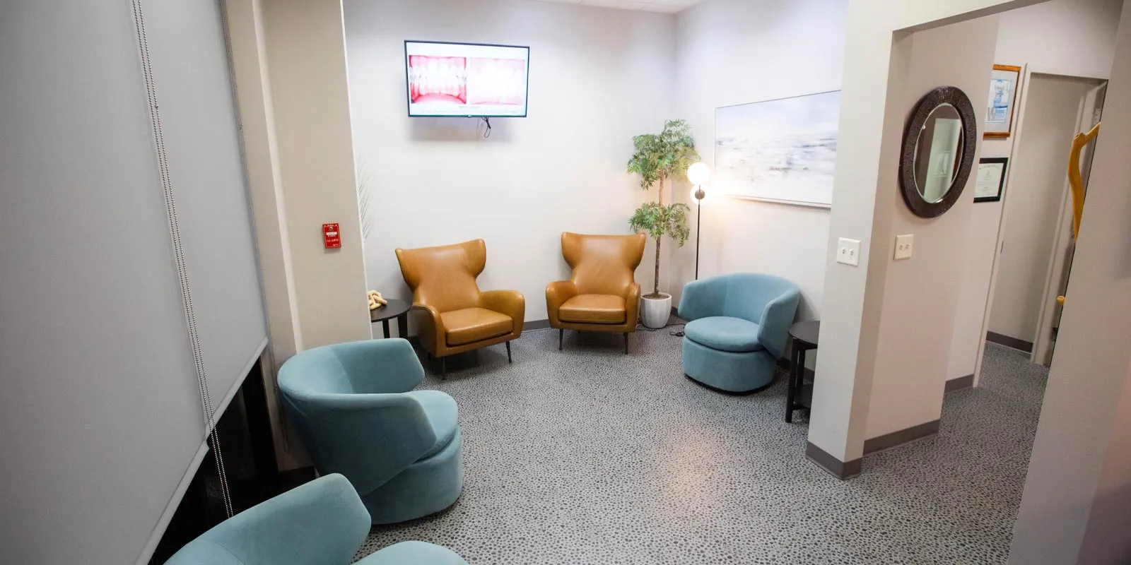 Modern waiting room with brown and blue chairs, a plant, floor lamp, and a wall-mounted TV screen.