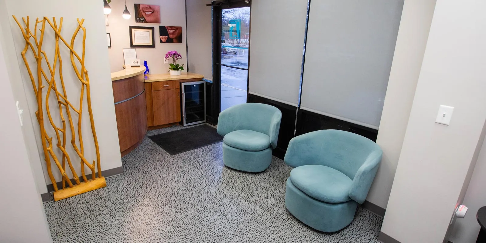 Modern waiting area with two blue chairs, wooden reception desk, decorative wooden sculpture, and window blinds.