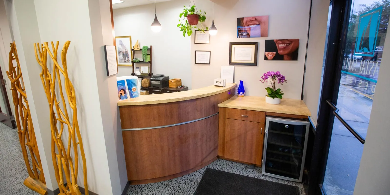 Reception area with curved wooden desk, plants, framed certificates, and a small refrigerator near glass entrance door.