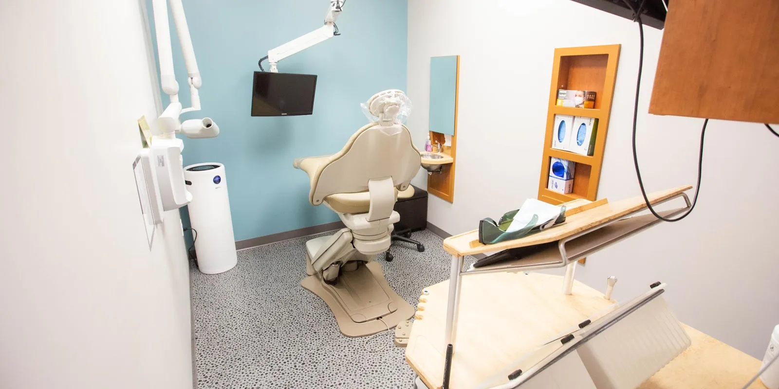 Modern dental clinic room with beige dental chair, overhead light, TV screen, and dental equipment.