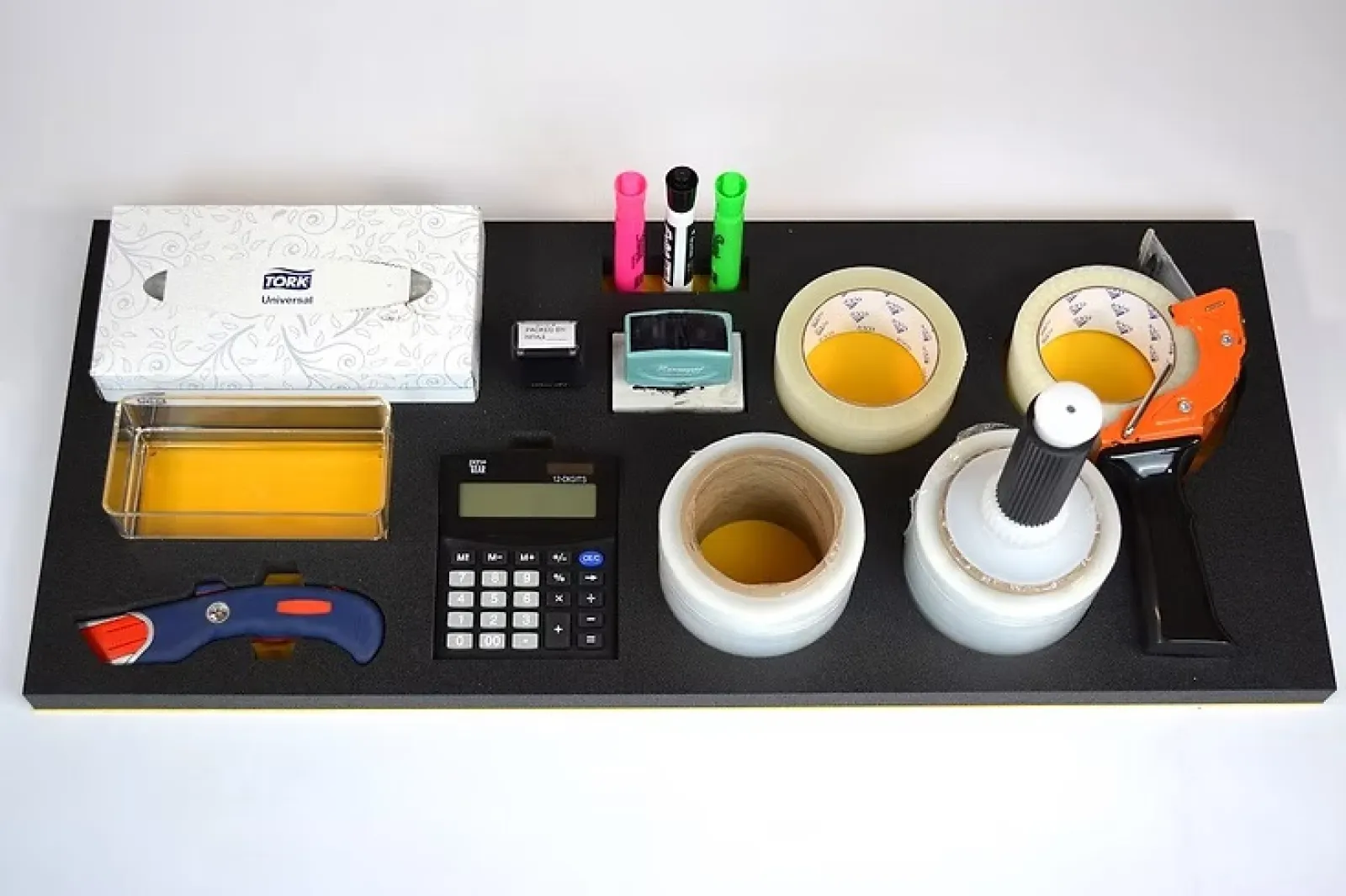 Organized office supplies including tissue box, highlighters, calculator, tape rolls, cutter, stamp, and tape dispenser on black board.