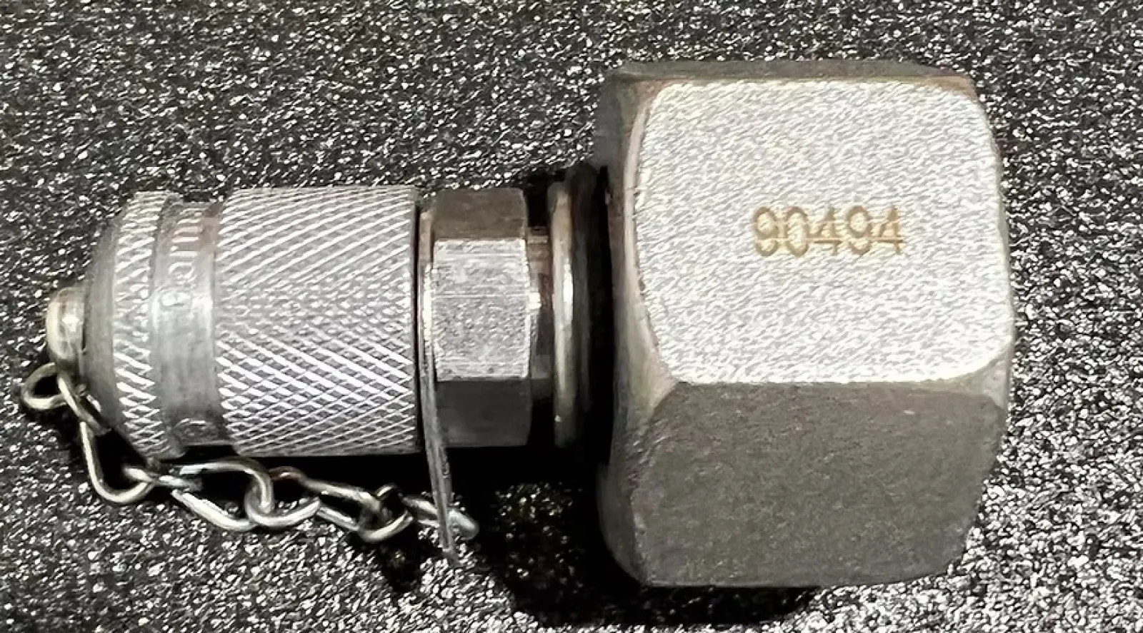 Metal hydraulic quick connect coupling with protective cap and chain on textured black surface.