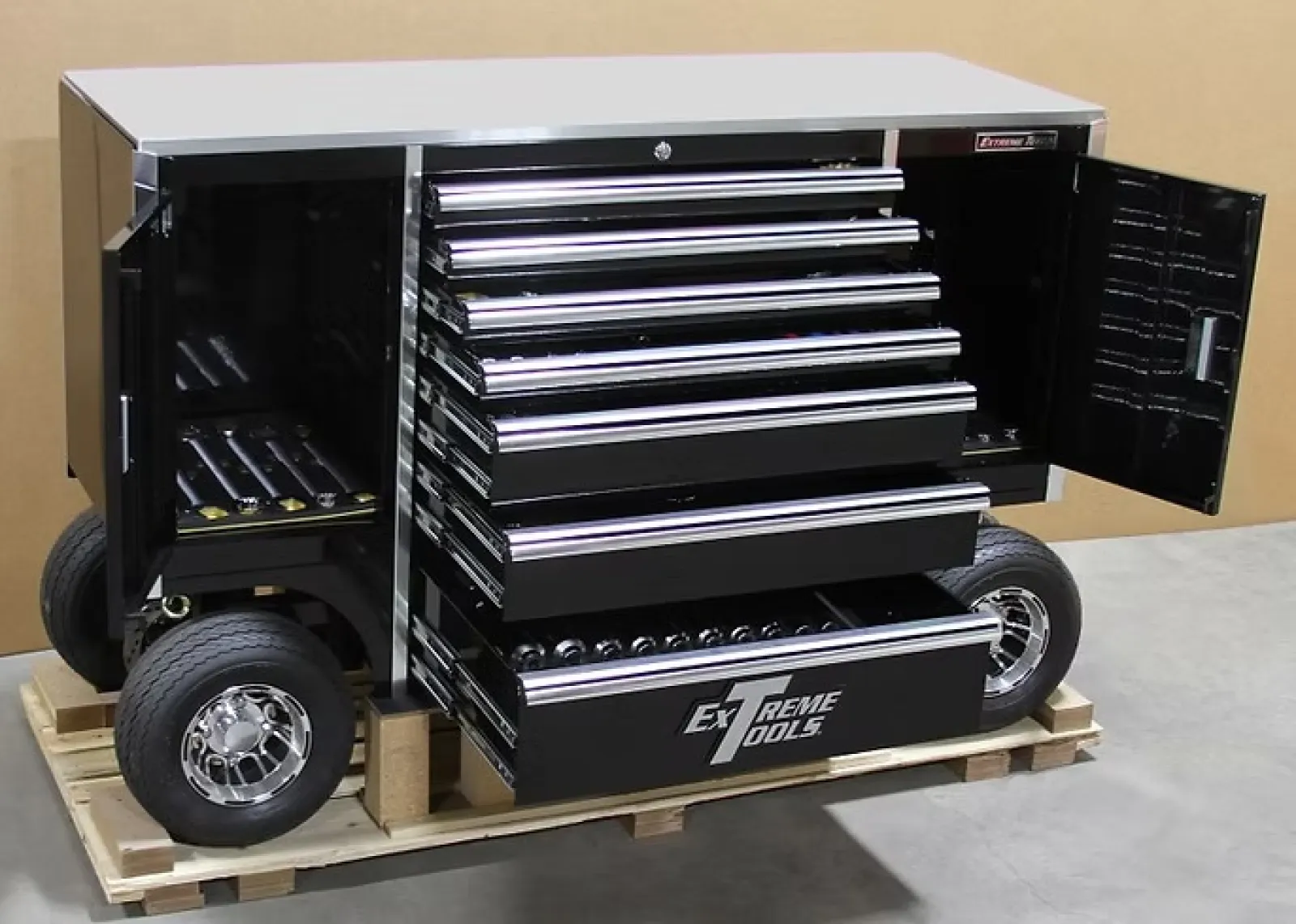 Black mobile tool chest with multiple open drawers and wheels on a wooden pallet indoors.