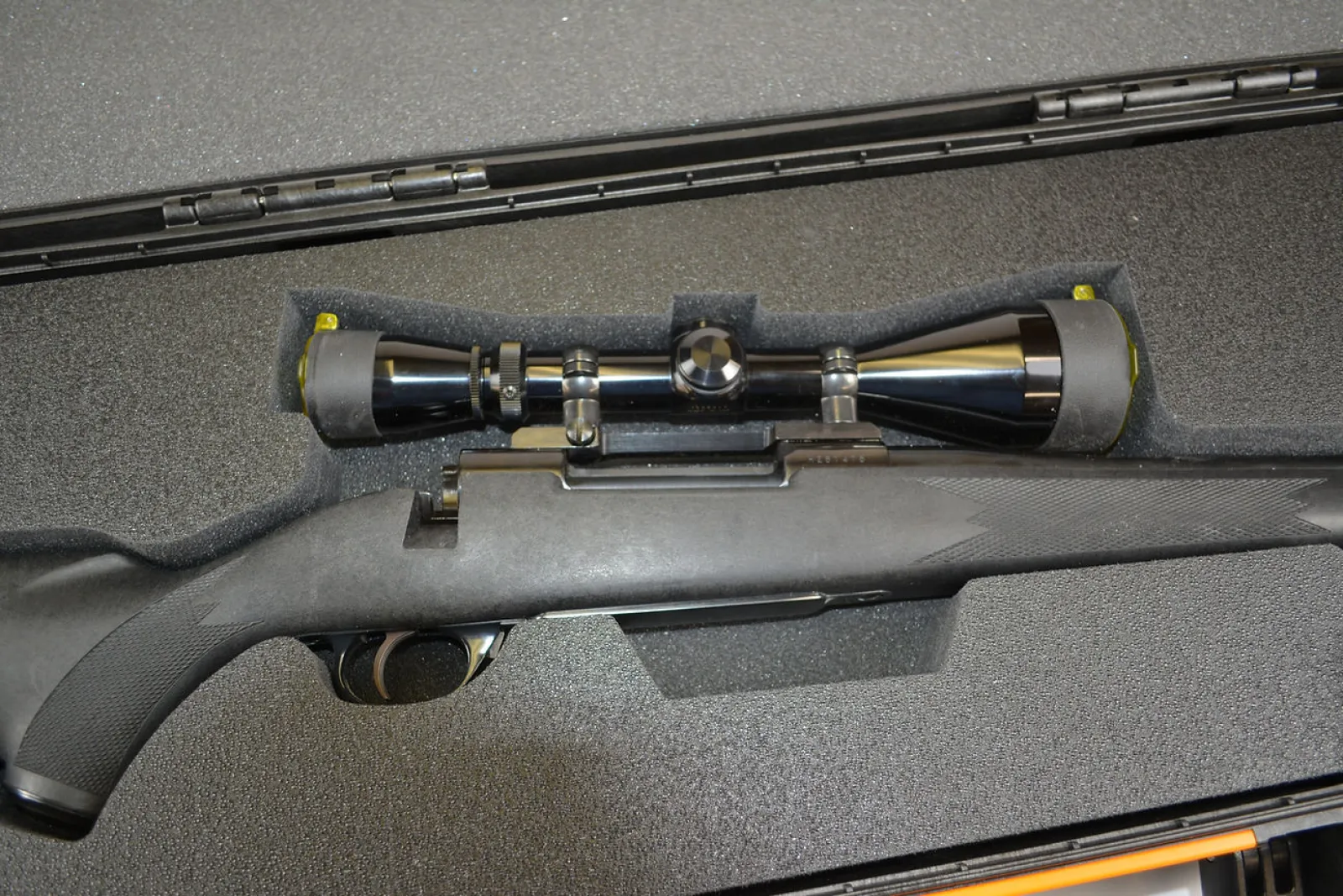 Black hunting rifle with a mounted scope inside a protective foam case on a textured surface.