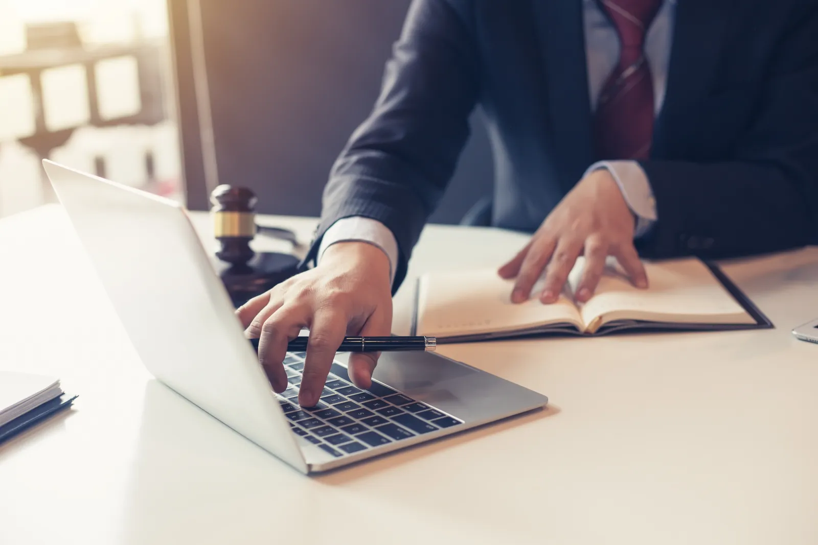 Business professional in suit working on laptop with open notebook and gavel on desk in office setting