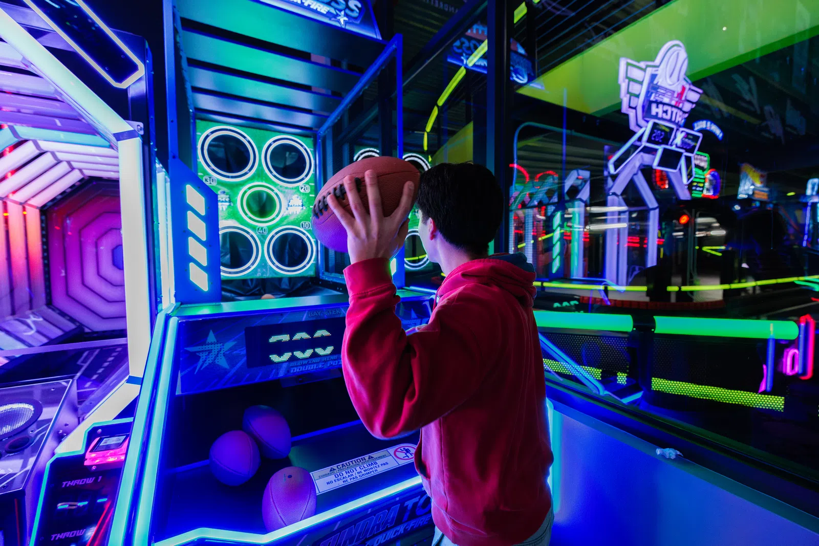 Person in red hoodie preparing to throw football in neon-lit arcade basketball game area