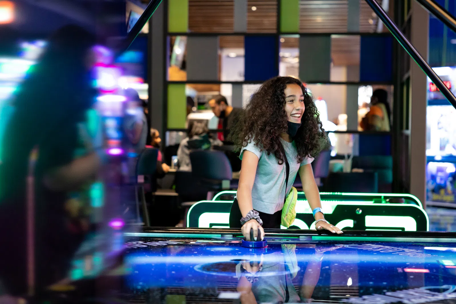 Happy girl playing air hockey in a vibrant arcade, surrounded by colorful lights and excited players.