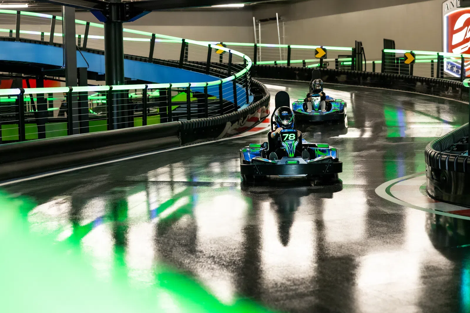Two racers driving green and black go-karts on an indoor electric kart track with neon green lights.