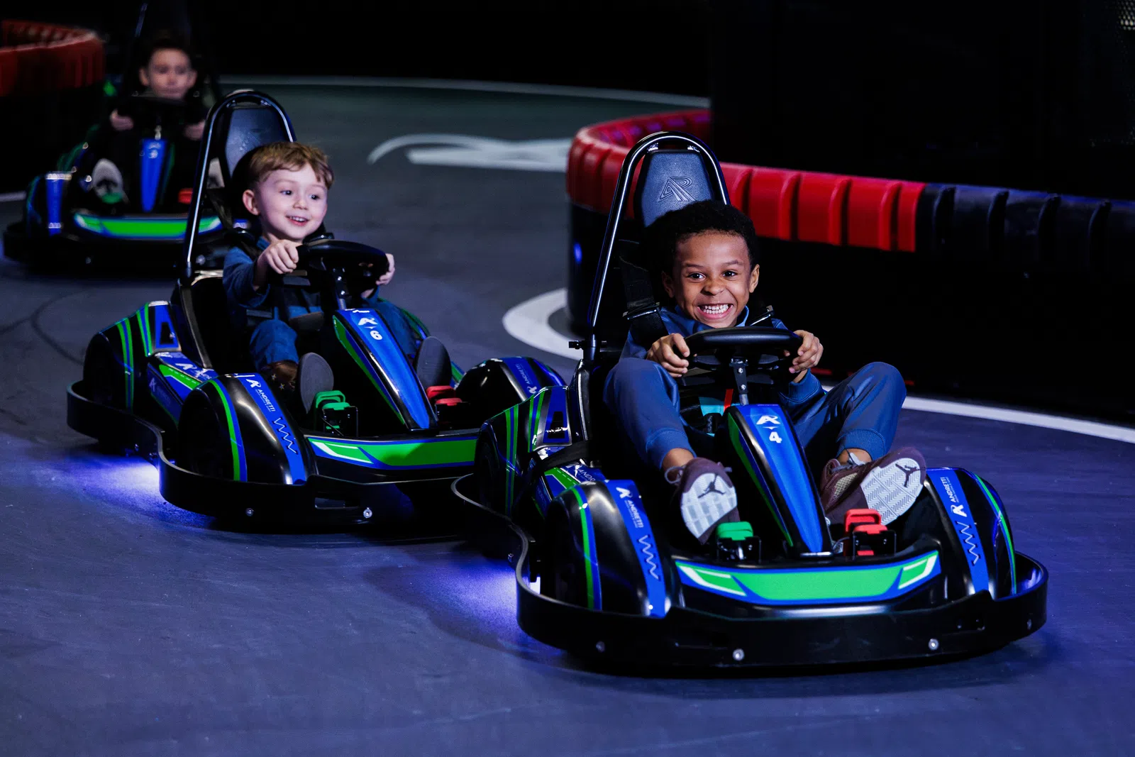 Two children smiling and racing in electric go-karts on an indoor track with glowing blue lights.