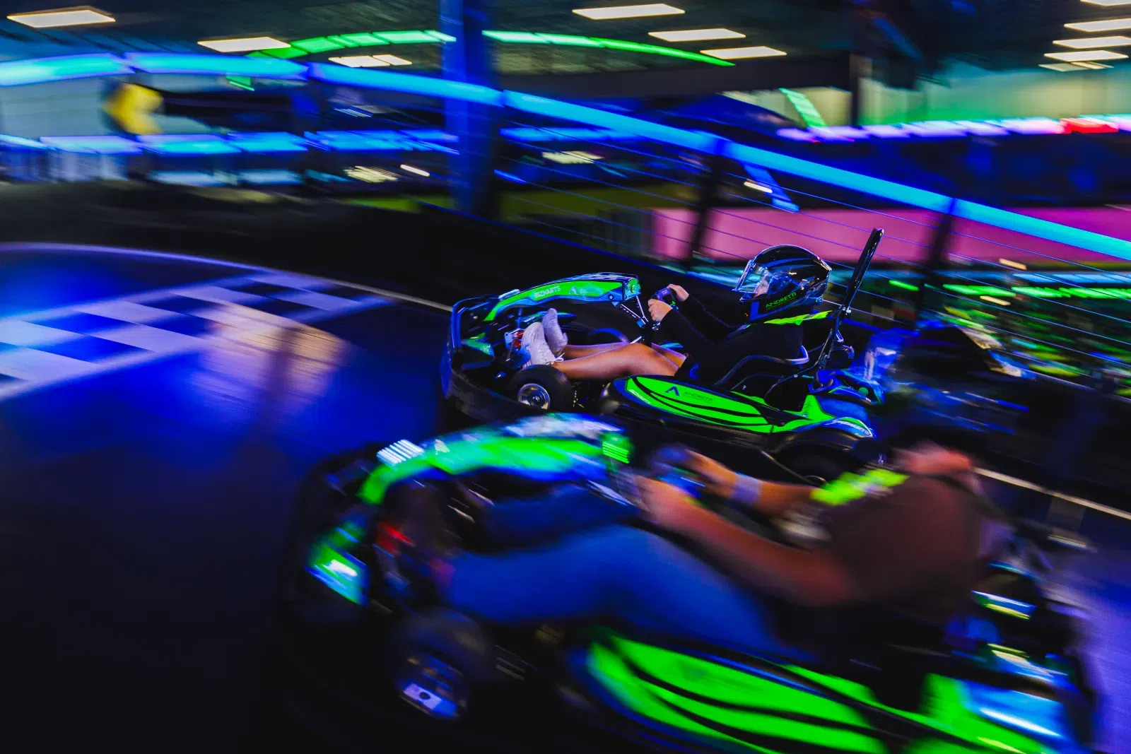 Two people racing go-karts on an indoor track with vibrant neon green and blue lights and motion blur