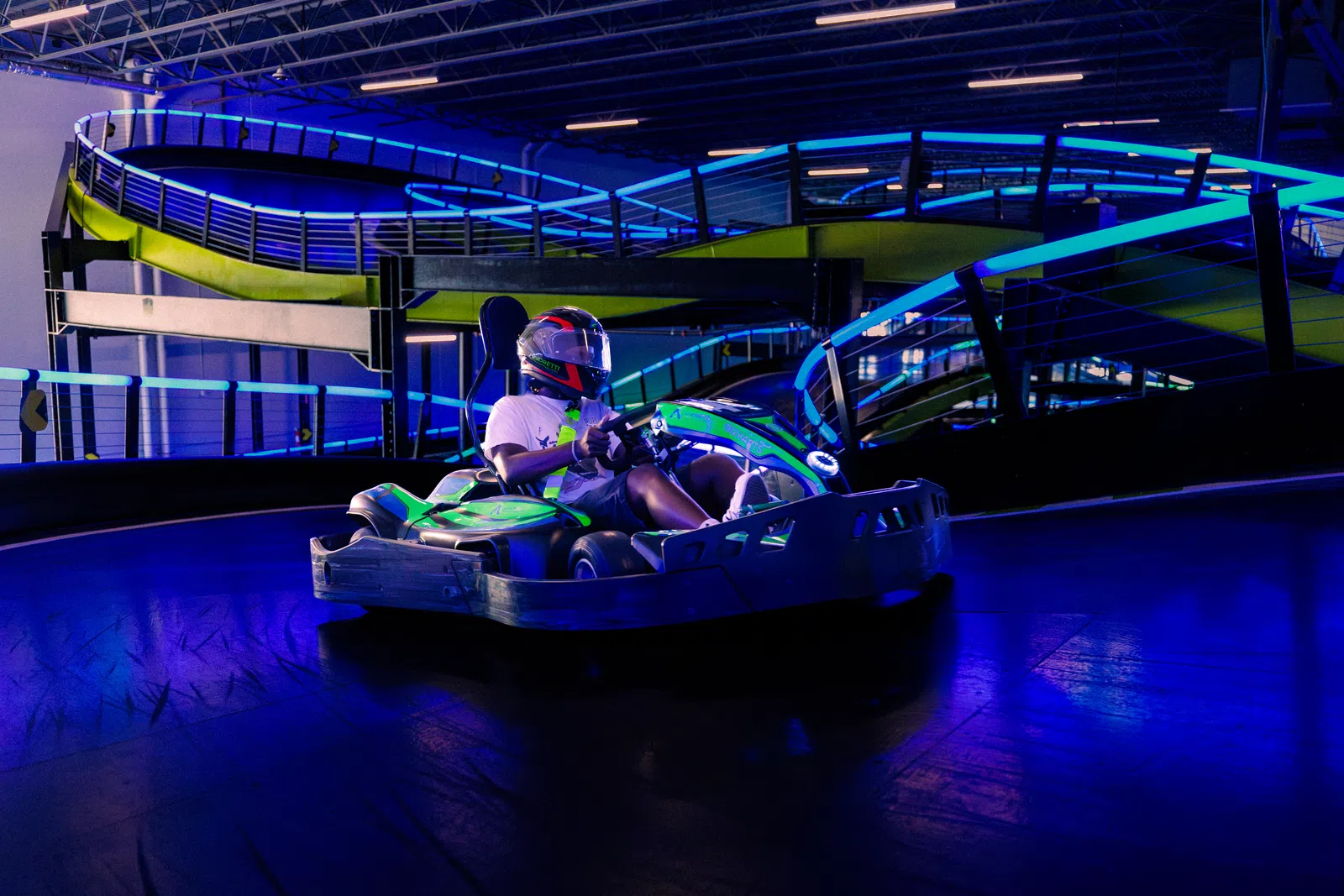 Person racing a green go-kart on an indoor track with blue and purple neon lights and safety railings.