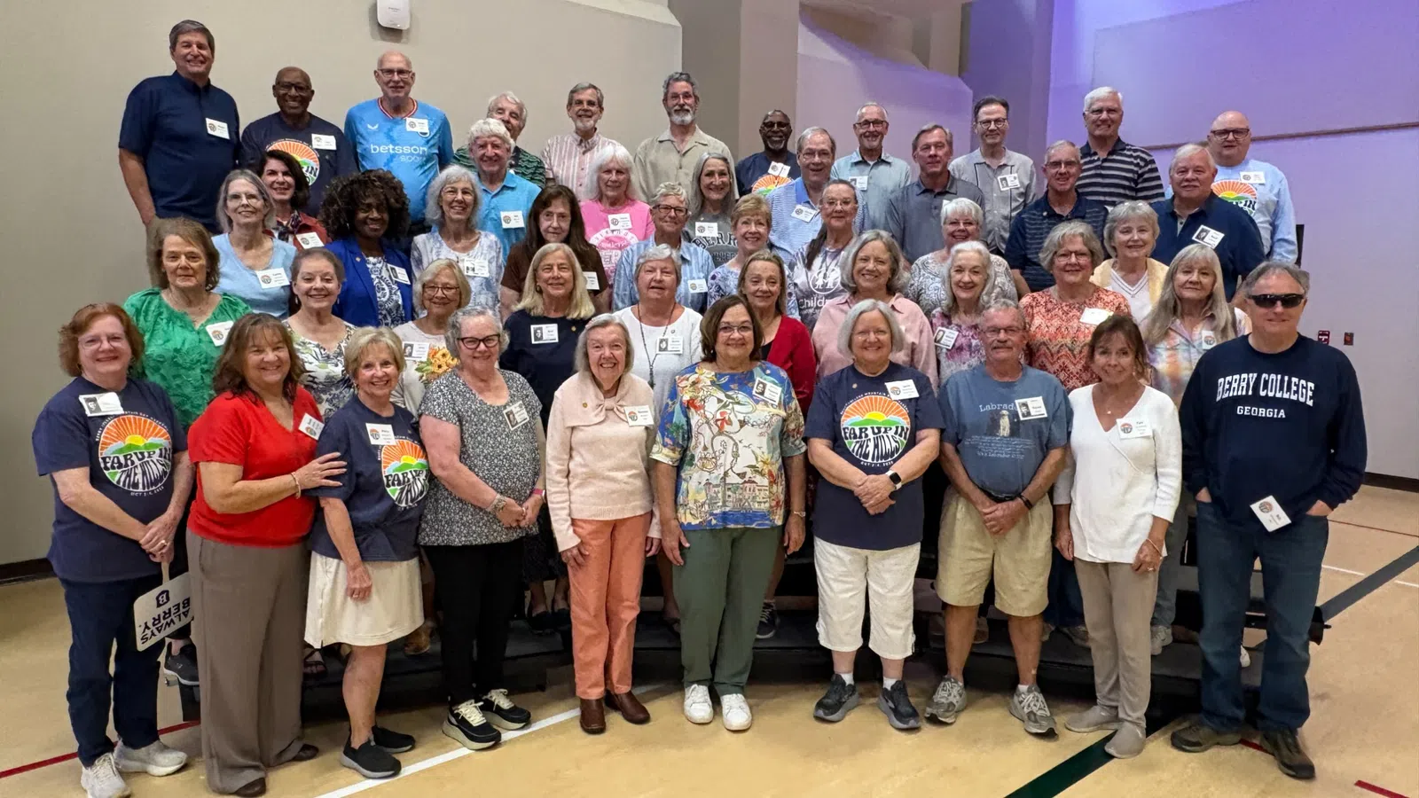 Large group of diverse adults smiling and posing indoors on gym floor with name tags, casual clothing, and bright lighting