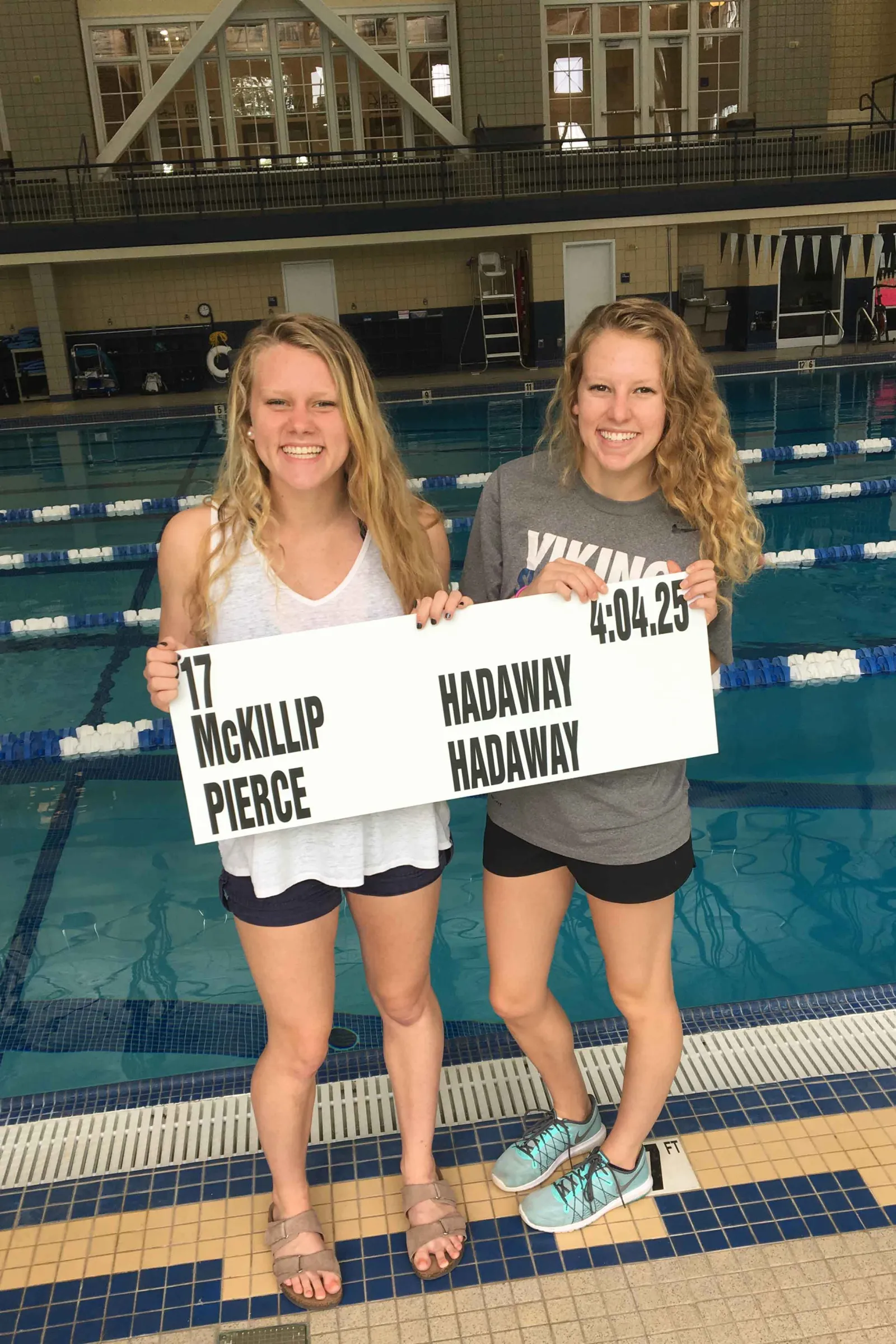 Two students stand in front of a pool holding a sign that reads &quot;17 McKillip Hadaway Pierce Hadaway 4:04.25&quot;