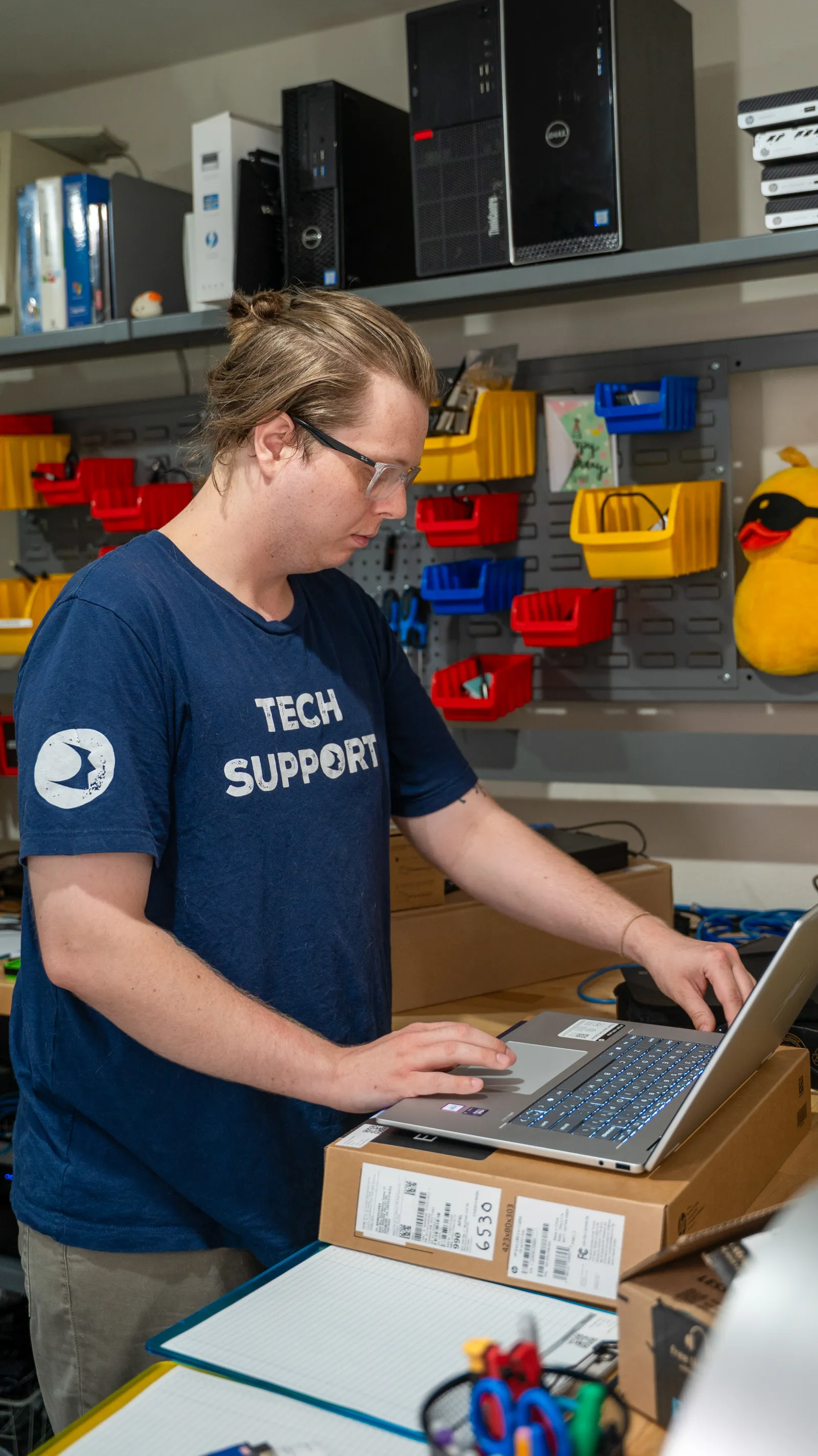 Tech support worker in blue shirt using laptop on a stack of boxes in a cluttered office workshop.