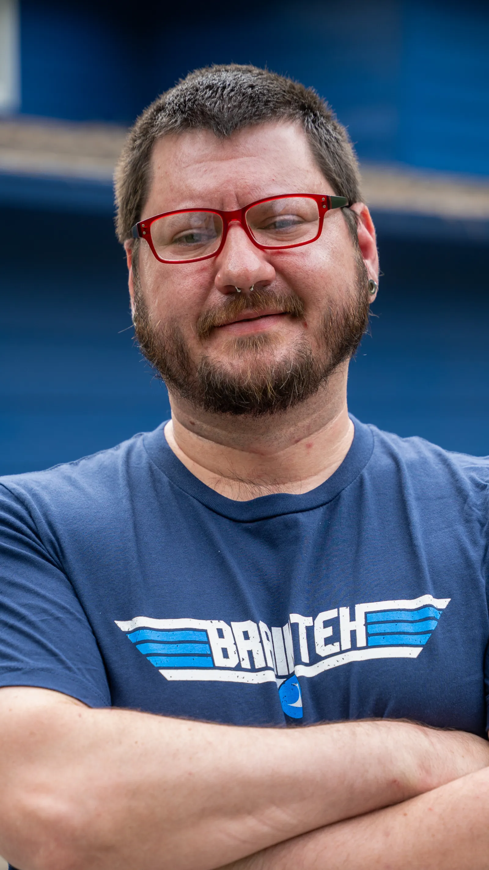 Bearded man with red glasses and septum piercing wearing a blue Brutech t-shirt with arms crossed.